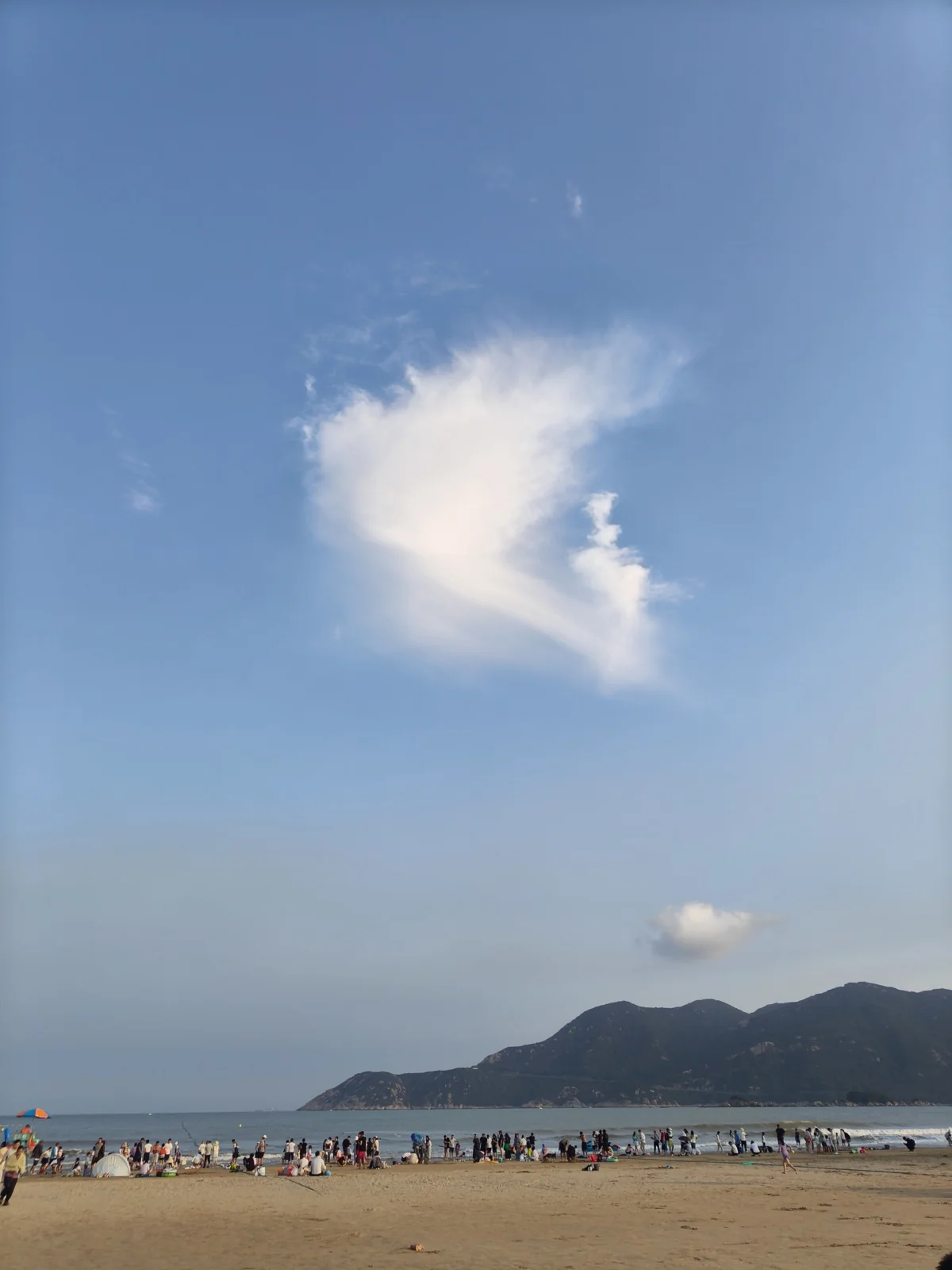 日落时分的南沙海滩，沙滩上聚集着许多游客，远处是山峦与蓝天白云