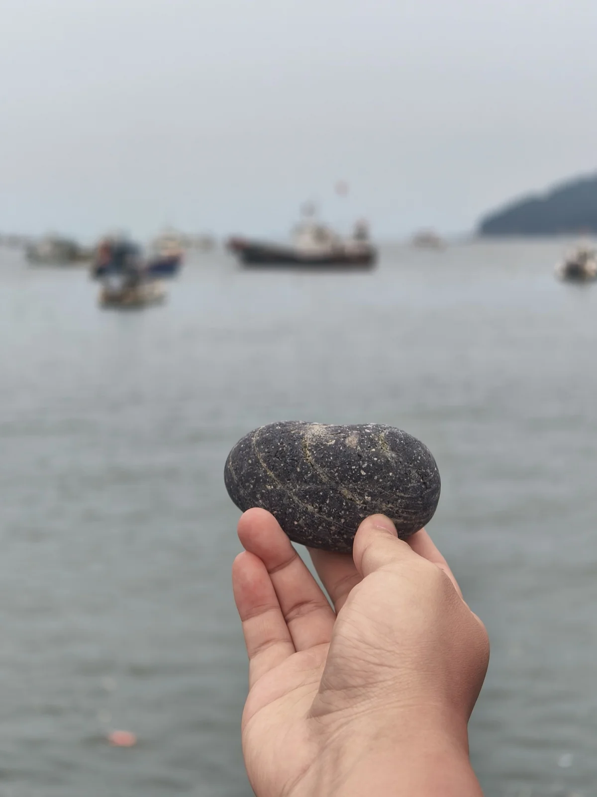 乌石塘海边，一只手握着一颗光滑的黑色鹅卵石，背景是模糊的海面与渔船