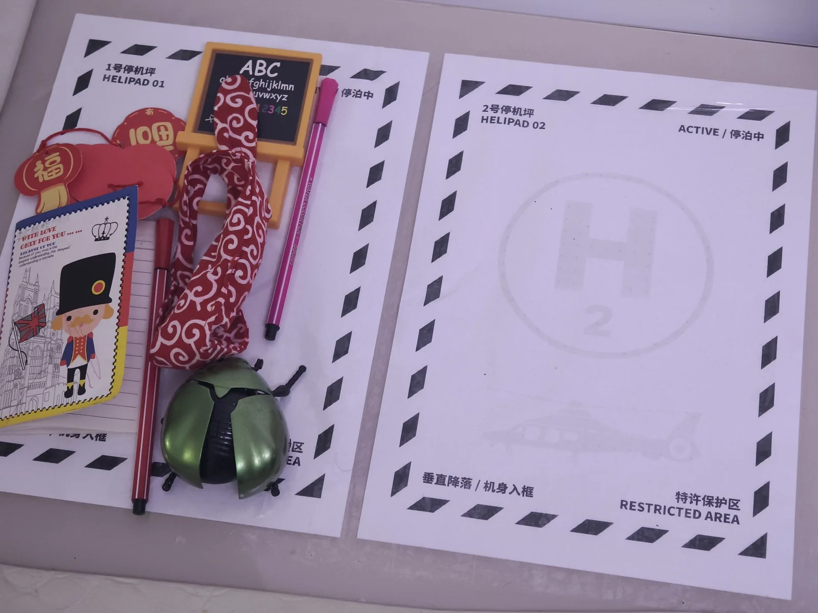Two printed A4 helipad sheets taped to a desk corner, Helipad 01 piled with a wooden alphabet board, festive red ornaments, a scarf, pens, and a green toy beetle, while Helipad 02 stays empty