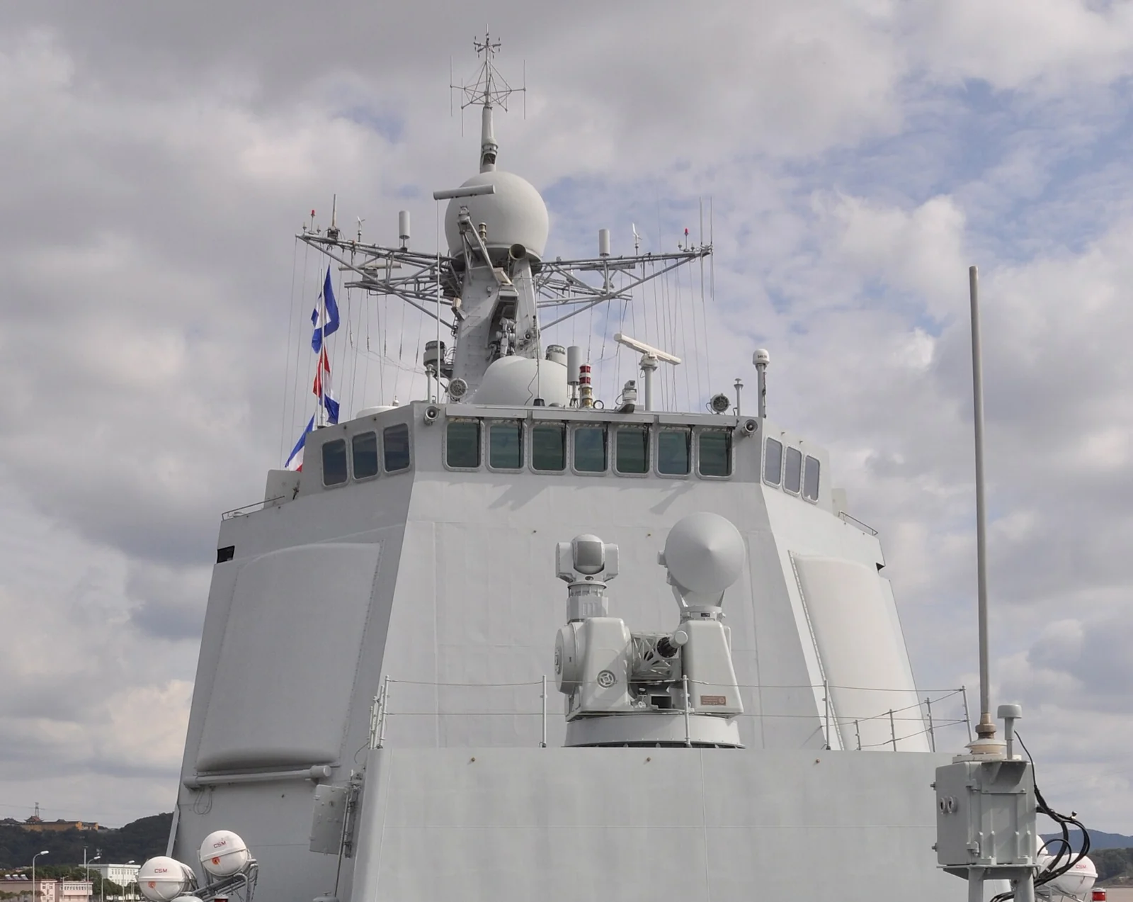 Close-up of a modern warship bridge and mast, with flat radar panels, a white radome, and a fire-control tracker.