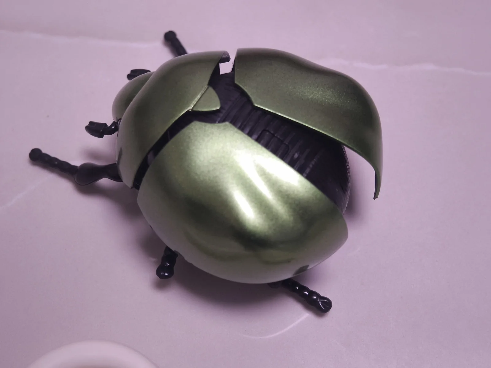 Green metallic toy beetle with its wing covers opened to expose the black body and legs, lying on a pale surface mid-repair