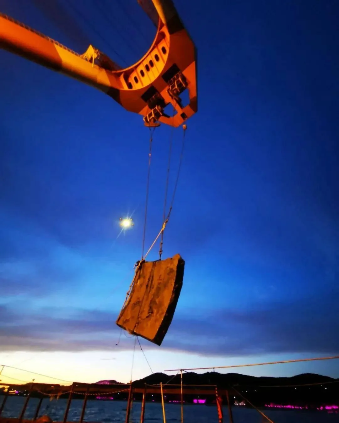 At dusk an orange crane lifts a thick dark slab over the water, lights along the distant shore.