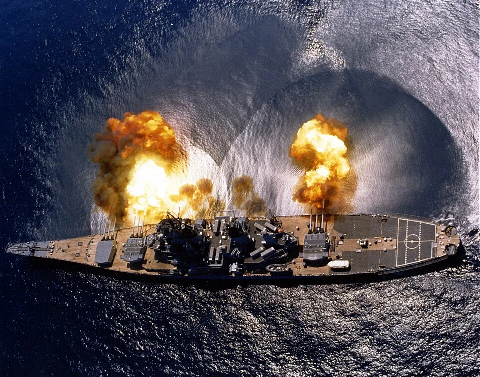Aerial view of an Iowa-class battleship firing a nine-gun starboard broadside, circular blast marks on the water.