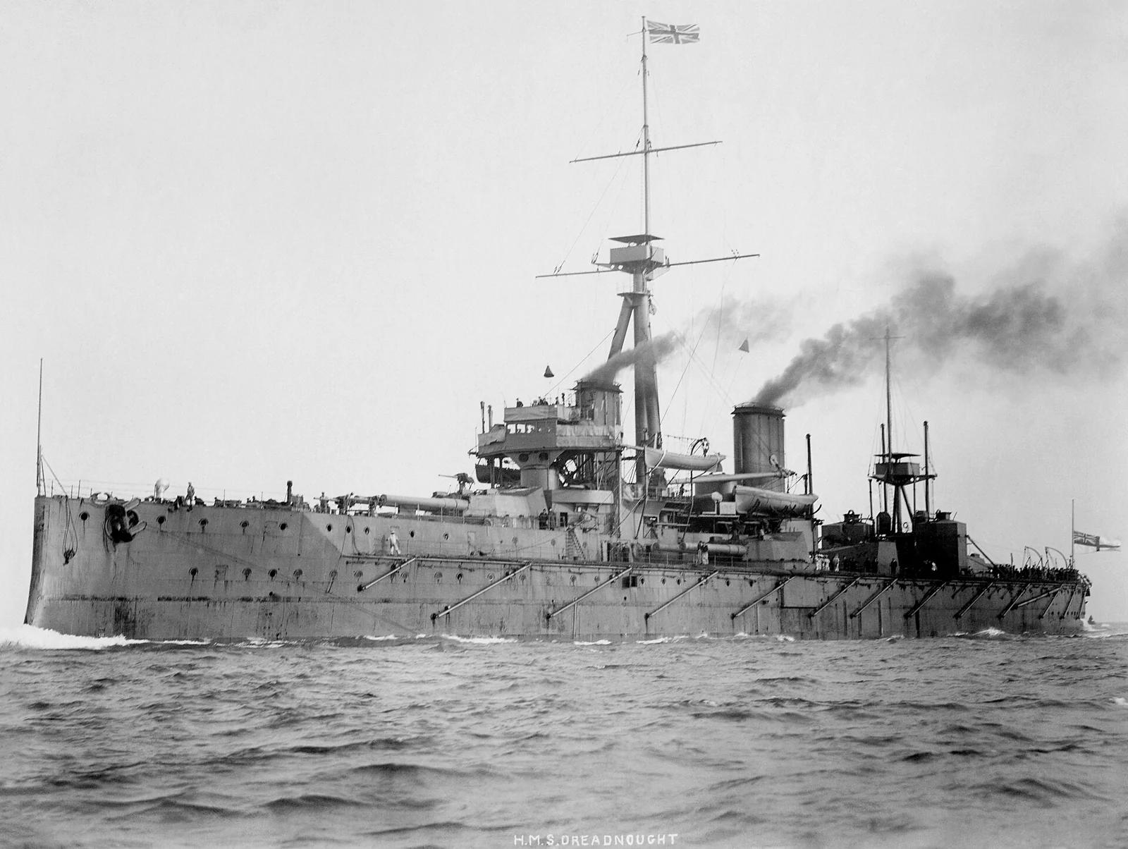 HMS Dreadnought underway, dark smoke from two funnels and several main turrets visible along the deck.