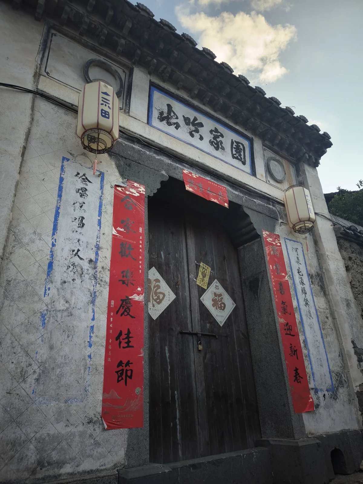 白墙老宅的深色木门前贴着红春联与褪色的蓝字旧联，门额题着山哈家园