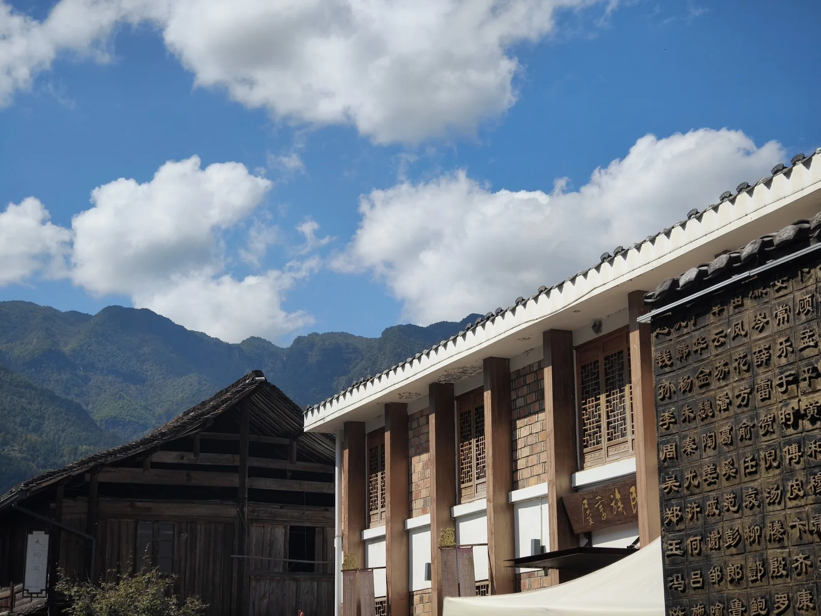 白墙青瓦的两层建筑与旁边深色木板老屋，右墙刻满汉字姓氏，背后是连绵青山