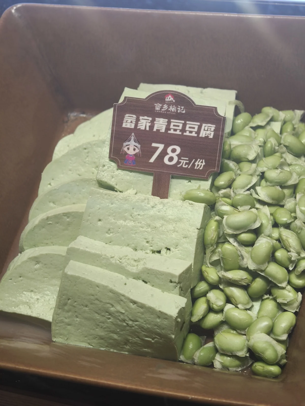 冷柜里摆着切片的青绿色豆腐和旁边的毛豆，插着畲家青豆豆腐的价格牌