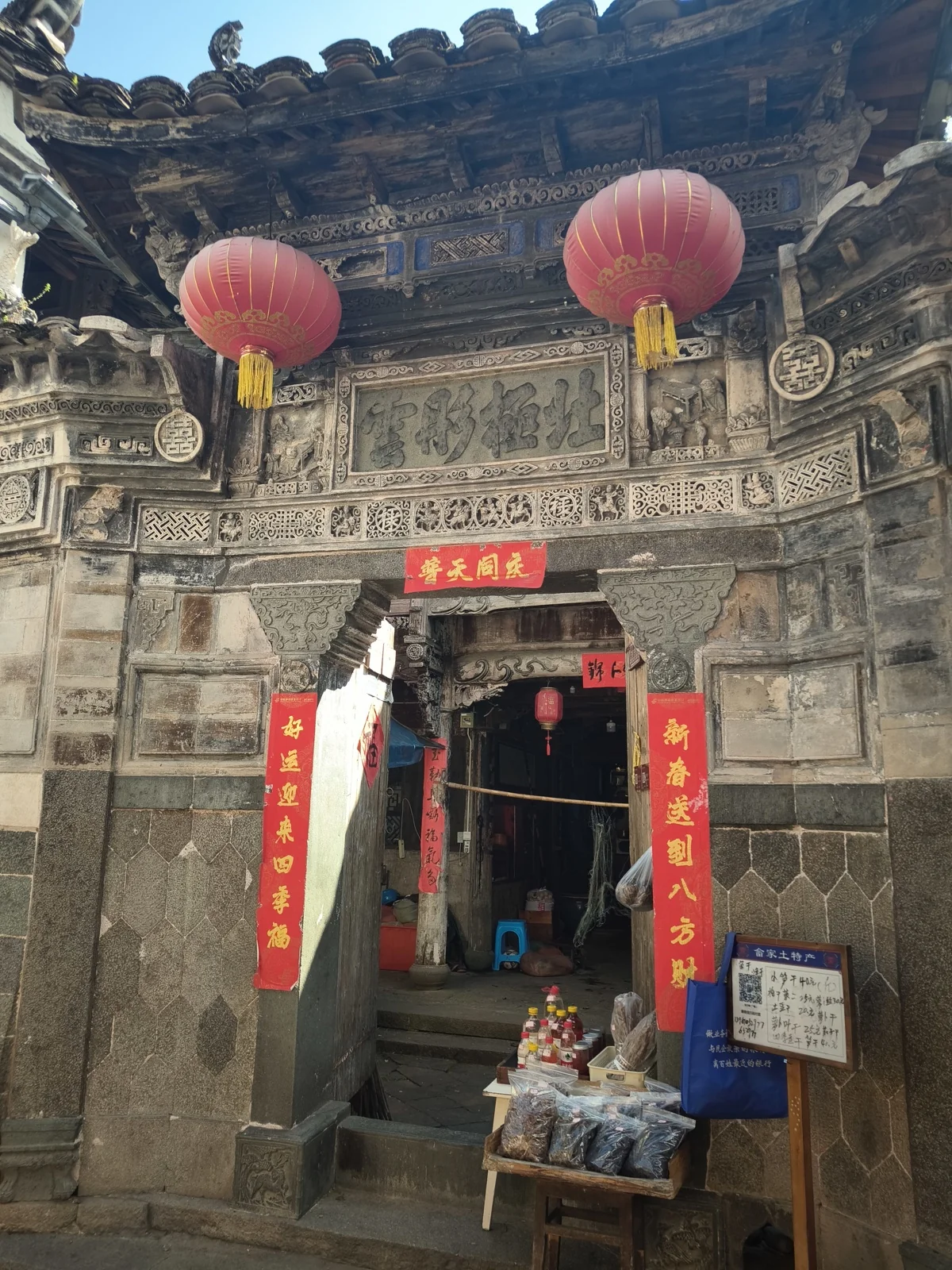 石雕门楼的老宅正门，砖雕门额下挂着红灯笼，门边摆着畲家土特产摊