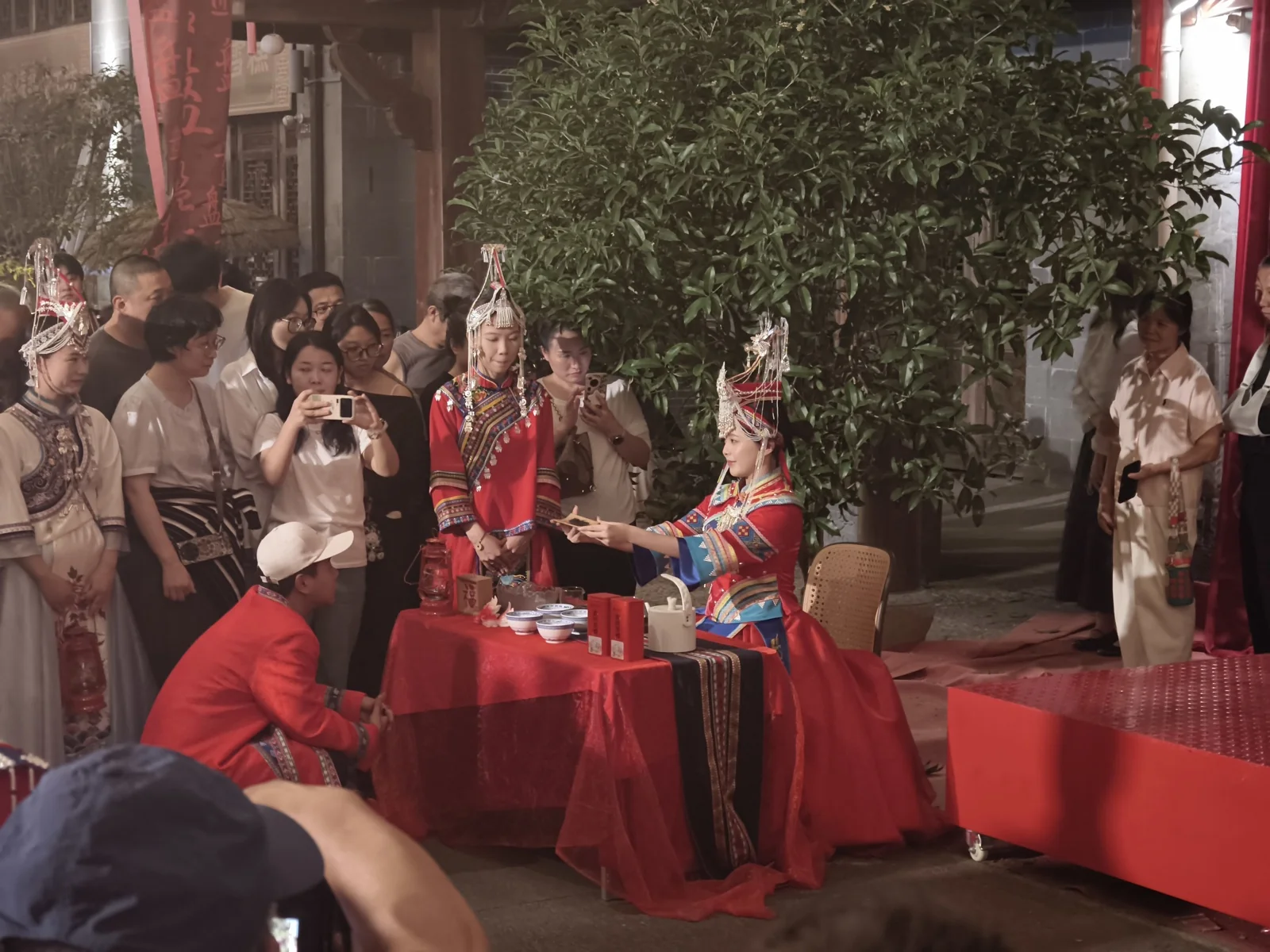 铺红布的桌前，穿红色婚服的女子向跪着的男子敬茶，旁有人举手机拍摄